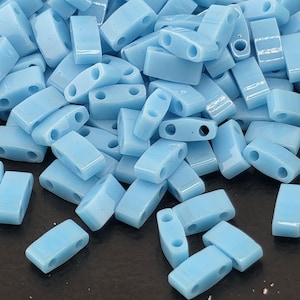May include: A pile of light blue rectangular beads with two holes each. The beads are smooth and glossy, with rounded edges. The beads are scattered, creating a textured surface.