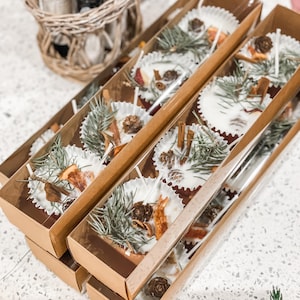 May include: Several white, round candles in clear boxes. Each candle is decorated with pine needles, dried orange slices, cinnamon sticks, and small pine cones. The candles are in brown cardboard boxes with clear plastic lids.