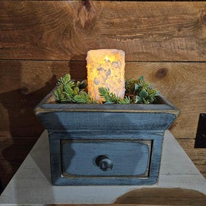 May include: A gray wooden box with a drawer and a candle inside. The box is sitting on a white wooden table. The candle is lit and has a white wax exterior.