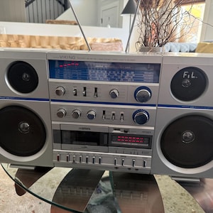 May include: A silver Viking portable stereo cassette recorder featuring a 3-way speaker system with four circular black speakers. The front panel includes a blue radio tuner display, multiple silver control knobs, sliding switches, and a cassette deck.
