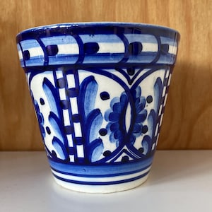 May include: A ceramic pot with a blue and white floral design. The pot features a repeating pattern of blue flowers and geometric shapes against a white background. The rim has a blue and white striped pattern. The pot is suitable for plants.