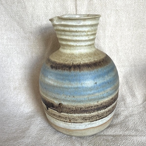 May include: A handmade ceramic jug with a rounded body and a spout. The jug features horizontal bands of colour, including light blue, brown, and beige. The top has a textured, ribbed design.