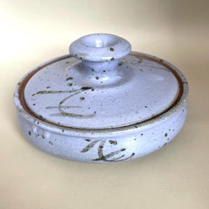 May include: A ceramic lidded container in a soft blue hue with speckled detailing. The lid features a small, rounded handle and a brown rim. The container has a matching base and is adorned with dark brown, abstract markings. A handmade pottery piece.