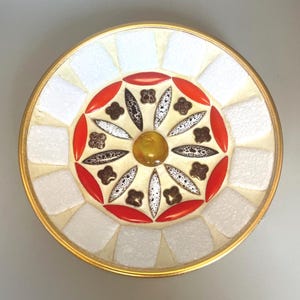 May include: A circular decorative dish with a mosaic design. The centre features a yellow glass piece surrounded by a red and brown floral pattern. The outer ring has a white and cream-coloured mosaic design, with a gold-coloured rim.