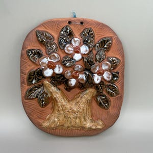 May include: A ceramic wall hanging depicting a tree with white flowers and brown leaves. The tree is set against a terracotta-coloured oval background. The piece has a rustic, handmade appearance and includes a blue string for hanging.