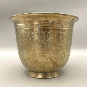 May include: A vintage brass planter with an etched floral and bird design. The planter has a wide, slightly flared rim and a rounded body. The brass has a warm, aged patina, and the design is intricate and detailed.