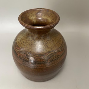 May include: A small, round ceramic vase with a wide opening. The vase has a speckled, earthy colour palette of brown, tan, and reddish-orange hues. The design includes horizontal bands of colour, adding visual interest to the piece.