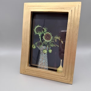 May include: A rectangular picture frame with a metallic gold finish containing a print of sunflowers in a textured blue and brown vase. The print features a dark background with green foliage and a small yellow bottle beside the vase.
