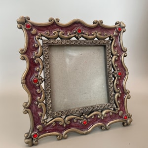May include: An ornate burgundy picture frame with gold-coloured accents and red jewel embellishments. The frame has a square opening and a decorative, scrolled design. A vintage-style home decor item.