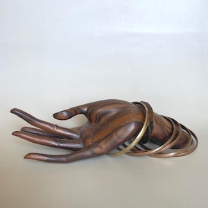 May include: A wooden hand sculpture with detailed grain patterns holds several thin metal bangles. The bangles are in shades of gold and copper. The sculpture is a warm brown colour and sits on a neutral background.