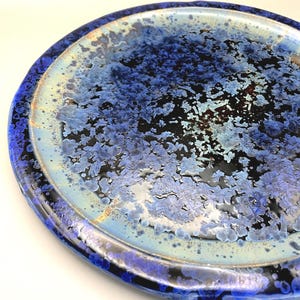 May include: A round ceramic plate with a glossy finish. The plate has a dark blue rim and a centre with a blend of blue, black, and white crystalline patterns. The design gives the plate a unique, textured appearance.
