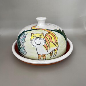 May include: A white ceramic butter dish with a domed lid. The lid features a hand-painted illustration of a yellow cat with orange stripes walking through a green field with flowers. The dish has a white rim and an orange base.