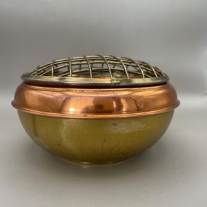 May include: A vintage brass and copper flower frog. The bowl-shaped container has a copper band around the middle and a brass base. A brass grid sits on top, designed to hold flower stems in place.