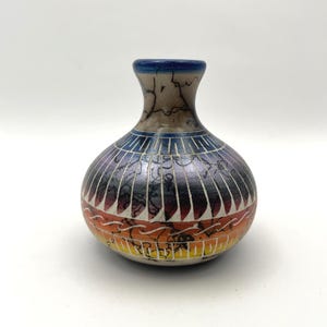May include: A small, round ceramic vase with a narrow neck. The vase has a colour gradient from orange to blue, with a detailed pattern of vertical lines and decorative elements. The rim is blue.