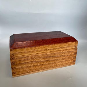 May include: A handcrafted wooden box with a deep reddish-brown lid and a lighter, natural wood body. The box has detailed joinery and a rectangular shape, displaying the natural wood grain and craftsmanship.