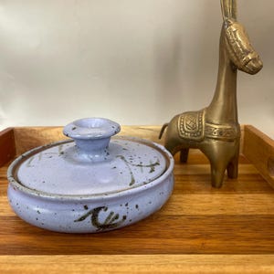 May include: A small, round, lavender ceramic dish with a lid, decorated with brown abstract designs. A brass donkey figurine stands nearby on a wooden tray. The dish has a small handle on the lid.