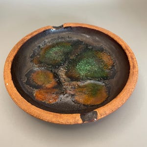 May include: A round, brown ceramic ashtray with a black and green glaze. The glaze has a mottled effect, with areas of dark brown, green, and orange.