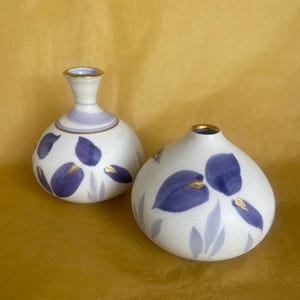 May include: Two white ceramic vases with a floral design. The vases have a bulbous shape and are decorated with purple and gold leaf accents. One vase has a narrow neck, while the other is round.