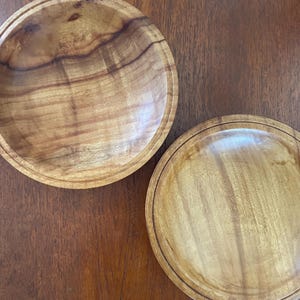 May include: Two wooden bowls with a natural wood grain pattern. The bowls are round with a slightly raised rim. The wood has a warm, light brown colour. The bowls are on a dark brown wooden surface.