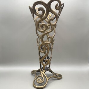 May include: A tall, open-work vase crafted from a metallic, bronze-toned material. The structure features an intricate, swirling pattern of interconnected metal loops and curves that form a hollow, trumpet-like shape with a wide, flared top.