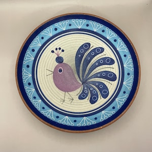 May include: Ceramic plate with a bird design. The plate has a blue and brown rim, with a light blue and white patterned border. The bird is purple with blue tail feathers and a crown. A decorative piece for the home, ideal for display.