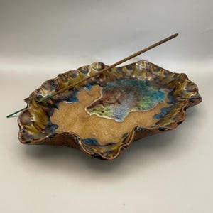 May include: A ceramic incense holder in a leaf shape. The holder has a tan base with blue, green, and brown accents. A brown incense stick is placed in the center. The edges are ruffled and have a dark brown glaze.