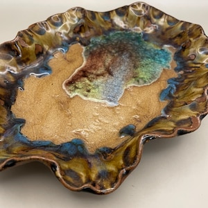 May include: A handmade ceramic leaf-shaped dish with a distinctive, organic design. The dish has a tan centre with a mottled pattern of brown, blue, and green around the edges. The rim has a wavy, textured appearance, adding to its artistic appeal.