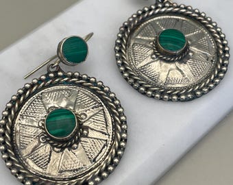 May include: A pair of circular silver earrings featuring a central malachite stone and a smaller malachite stud at the top. The silver discs have a textured, geometric pattern with a beaded border and a twisted rope edge surrounding the center stone.