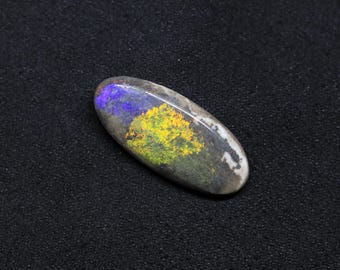May include: An oval-shaped opal gemstone with a polished surface. The opal displays a vibrant play of color, including blue, yellow, and green hues. The stone is set against a dark background, highlighting its iridescent qualities.