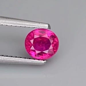 Puede incluir: Una gema redonda, facetada, de color rosa intenso, sujeta con unas pinzas plateadas. La gema tiene un tallado brillante con múltiples facetas. El fondo es gris claro.