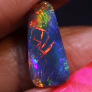 Könnte beinhalten: Ein polierter, länglicher Opal-Edelstein. Der Opal zeigt eine dunkelblaue Grundfarbe mit lebendigem internen Farbspiel, einschließlich Blitzen von Rot, Orange, Grün und Lila. Der Edelstein wird zwischen Fingern gehalten.