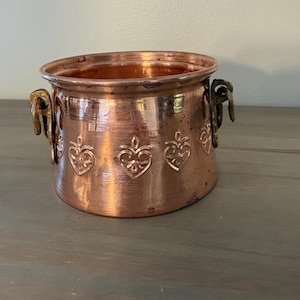 May include: A round copper pot featuring a polished metallic finish and a series of embossed heart-shaped motifs around the exterior. The vessel includes two dark, weathered metal handles attached to opposite sides of the upper rim.