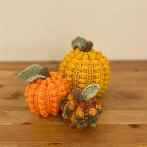 May include: Three crocheted pumpkins in different sizes and colors. The largest pumpkin is yellow, the middle pumpkin is orange, and the smallest pumpkin is a mix of orange, green, and brown.