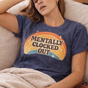 May include: A navy blue t-shirt with the text "MENTALLY CLOCKED OUT" in a retro rainbow design. The shirt is worn by a person resting on a couch.