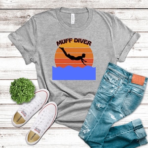 May include: A gray t-shirt with the text "MUFF DIVER" above a silhouette of a person diving into water, set against an orange and yellow sunset. The shirt is paired with blue jeans and white sneakers.