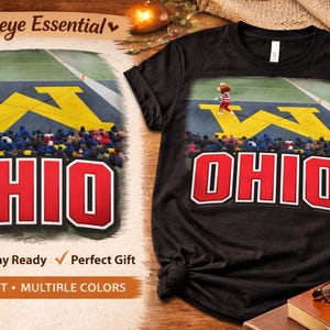 May include: Black t-shirt featuring a graphic of a football field with the word "OHIO" in red block letters. The image includes a cartoon figure and the text "Buckeye Essential".