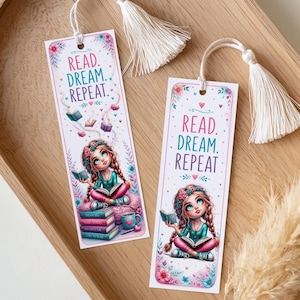 Könnte beinhalten: Zwei dekorative Lesezeichen mit weißen Quasten. Jedes Lesezeichen zeigt ein junges Mädchen beim Lesen, umgeben von Büchern und Blumen. Der Text "READ. DREAM. REPEAT." ist oben auf jedem Lesezeichen aufgedruckt.