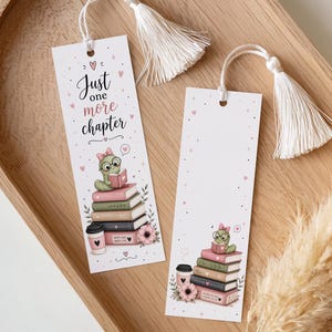 Könnte beinhalten: Zwei weiße Lesezeichen mit cremefarbenen Quasten liegen auf einem Holztablett. Ein Lesezeichen zeigt einen Cartoon-Wurm, der auf Büchern liest, mit dem Text "Just one more chapter". Das andere Lesezeichen ist leer.