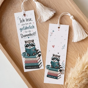 Può includere: Due segnalibri bianchi con nappe, raffiguranti un procione che legge un libro. I segnalibri hanno il testo "Ich lese, also bin ich gefährlich für die Dummheit!" e sono decorati con cuori e foglie. Il procione indossa gli occhiali e siede su una pila di libri.