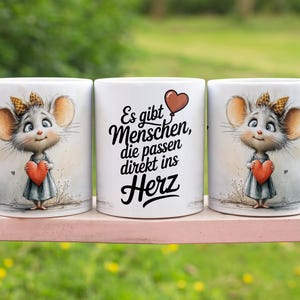 Könnte beinhalten: Drei weiße Keramikbecher. Zwei Becher zeigen eine Cartoon-Maus, die ein rotes Herz hält. Die Maus hat große Ohren und trägt ein Kleid. Der mittlere Becher hat deutschen Text und eine Herzgrafik.