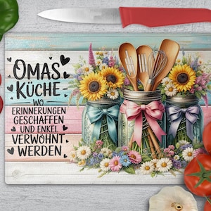 Op de afbeelding: Decoratieve snijplank met een bloemenontwerp met zonnebloemen en weckpotten. De potten zijn gevuld met bloemen en houten keukengerei. De plank heeft tekst in het Duits en wordt vergezeld door een rood mes en verse groenten.