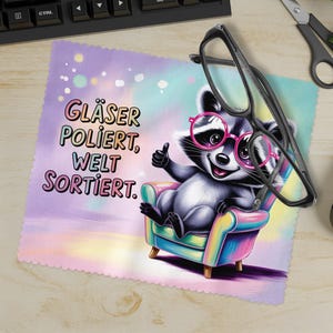 Pode incluir: Um pano de limpeza de microfibra com um guaxinim de desenho animado usando óculos rosa, sentado em uma poltrona colorida. O pano tem o texto "Gläser Poliert, Welt Sortiert." Óculos pretos estão sobre o pano.