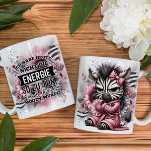 Könnte beinhalten: Zwei weiße Keramikbecher mit unterschiedlichen Designs. Ein Becher zeigt ein Zebra, das einen rosa Bademantel trägt, mit dem Text "Ich habe heute nicht die Energie so zu tun, als ob ich dich  morgen wurde!" Der andere Becher zeigt ein Zebra, das einen rosa Bademantel trägt, mit Blumen und Streifen im Hintergrund.