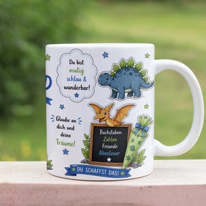 May include: White ceramic mug featuring cartoon dinosaurs and text in German. The mug has a blue Stegosaurus, a Pterodactyl, and a chalkboard with words. The mug also has text that says "Du bist mutig schlau & wunderbar!" and "DU SCHAFFST DAS!"