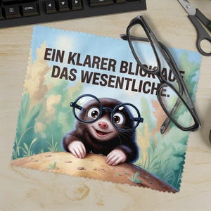 May include: A light blue cleaning cloth with a cartoon mole wearing glasses. The cloth has the German text "EIN KLARER BLICK AUF DAS WESENTLICHE." A pair of black glasses and scissors are on the wooden surface.
