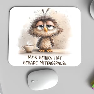 Op de afbeelding: Een witte muismat met een cartoon uil, een kopje koffie en de Duitse tekst "MEIN GEHIRN HAT GERADE MITTAGSPAUSE". De uil heeft slaperige ogen. Een computermuis, toetsenbord en koffiekopje zijn ook zichtbaar.