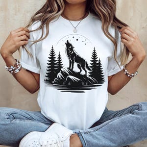Puede incluir: Camiseta blanca con un diseño en blanco y negro de un lobo aullando sobre una roca, con pinos y un cielo estrellado de fondo. El diseño está centrado en la parte delantera de la camiseta.