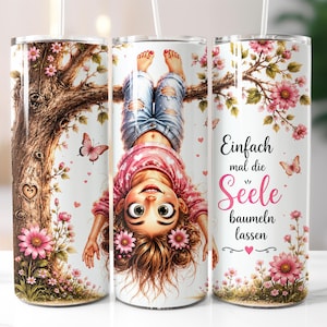 Könnte beinhalten: Ein zylindrischer Becher mit einer Grafik einer Person, die kopfüber an einem Ast hängt, umgeben von rosa Blumen und Schmetterlingen. Der Text lautet "Einfach mal die Seele baumeln lassen", ergänzt durch ein kleines Herzsymbol unter dem Text.