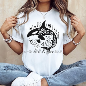 May include: White t-shirt featuring a black and white orca whale design with floral accents. The words "wild & free" are printed in a decorative font below the whale. The shirt is a classic crew neck style.