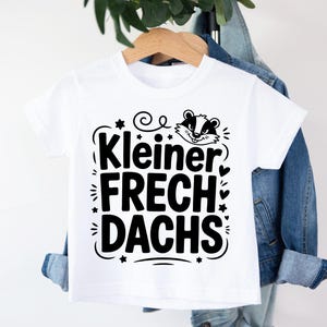Puede incluir: Camiseta blanca de manga corta con el texto negro "Kleiner Frechdachs", con una ilustración de tejón. El diseño incluye estrellas, corazones y espirales decorativas. La camiseta está colgada de una percha de madera.